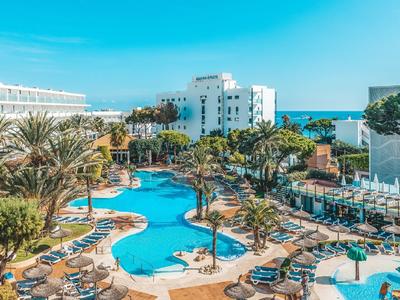 Ośrodek z dużym basenem, otoczony palmami, leżakami i białymi budynkami pod czystym niebem.