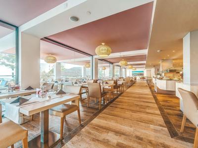 Przestronna restauracja hotelowa z drewnianą podłogą, beżowymi krzesłami i dużymi oknami wpuszczającymi światło dzienne.