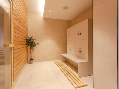 Heller Saunaraum mit Holzbänken, Pflanze in Ecke und Sauna aus hellem Holz links.