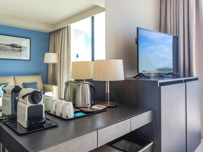 Moderner Hotelzimmerbereich mit Kaffeemaschine, Lampen, Fernseher und hellen Vorhängen am Fenster.