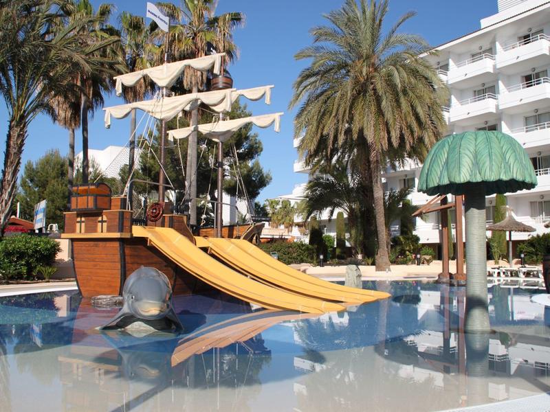 Strefa basenowa z zjeżdżalnią w kształcie statku pirackiego, palmami i hotelem w tle.