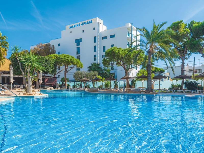 Hotel z dużym niebieskim basenem, palmami i leżakami pod czystym niebem.