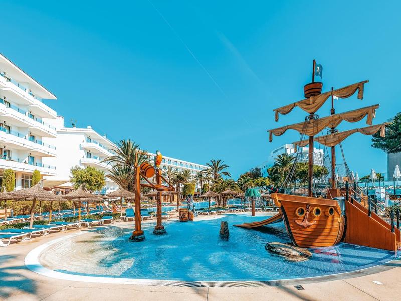 Basen w kurorcie z palmami, leżakami i placem zabaw w kształcie statku pirackiego pod niebieskim niebem.
