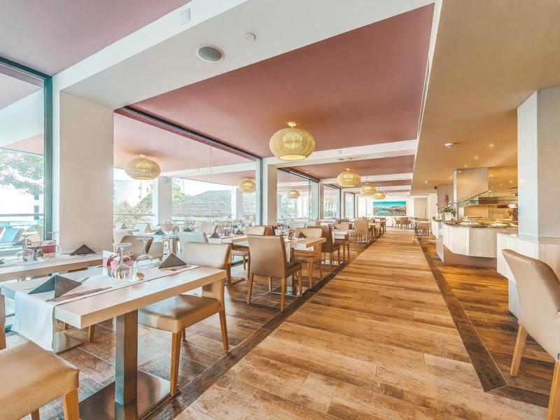 Przestronna restauracja hotelowa z drewnianą podłogą, beżowymi krzesłami i dużymi oknami wpuszczającymi światło dzienne.