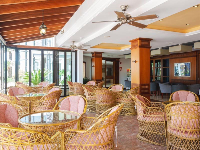 Jasny salon hotelowy z wiklinowymi krzesłami, szklanymi stolikami i wentylatorami sufitowymi pod drewnianymi belkami.