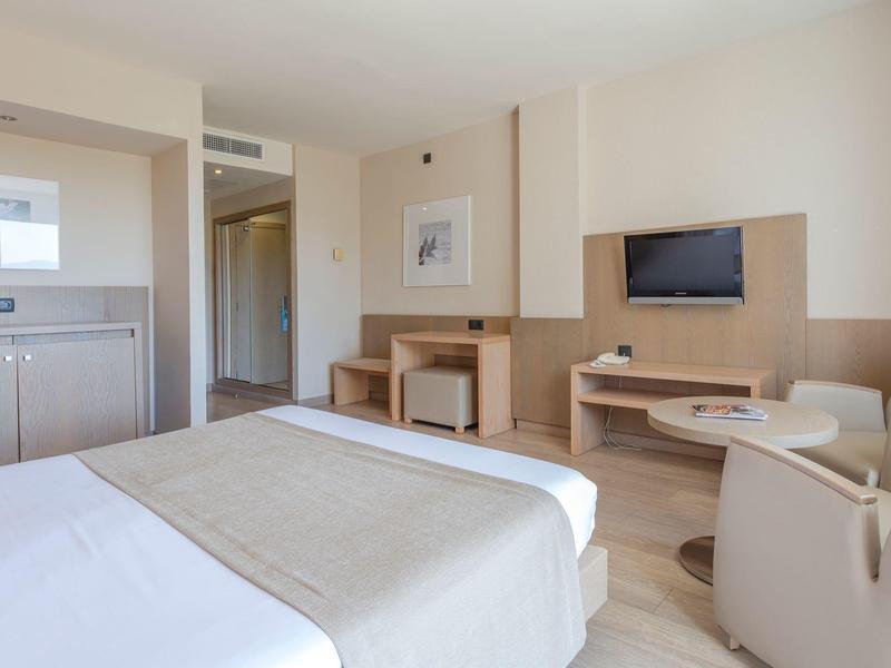 Nowoczesny pokój hotelowy z łóżkiem, fotelami, telewizorem, biurkiem i aneksem kuchennym w neutralnych kolorach.