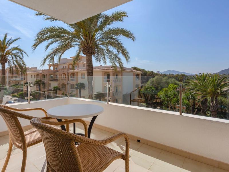 Balkon na zewnątrz z dwoma wiklinowymi krzesłami i okrągłym stołem z widokiem na palmy i budynki.