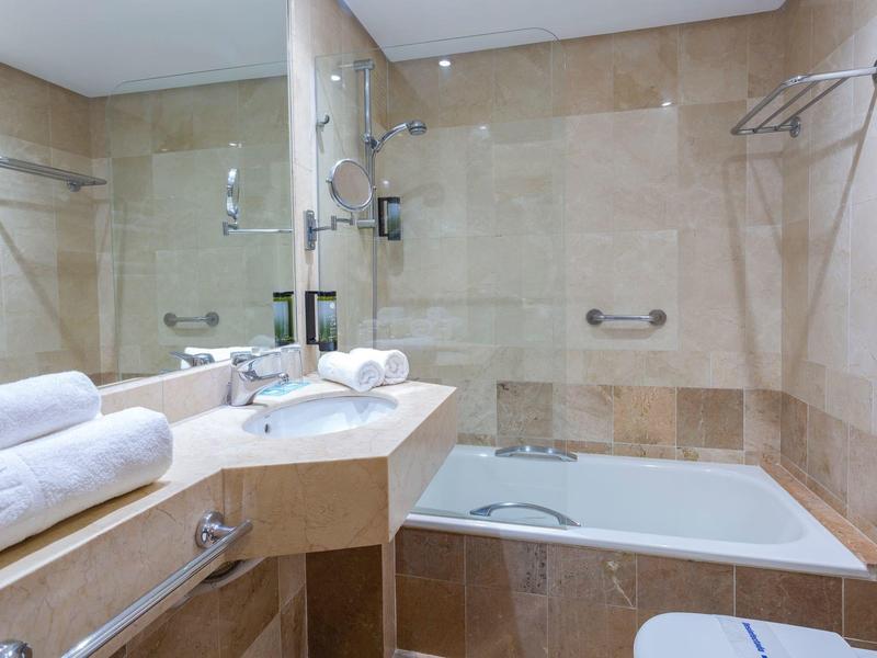 Nowoczesna łazienka z beżowymi płytkami, wanną, dużym lustrem i starannie złożonymi białymi ręcznikami.