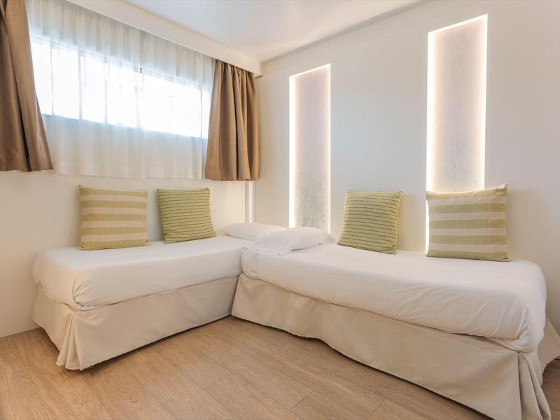 Jasny narożnik pokoju hotelowego z białymi sofami, zielonymi poduszkami, drewnianą podłogą i podświetlanymi panelami ściennymi.