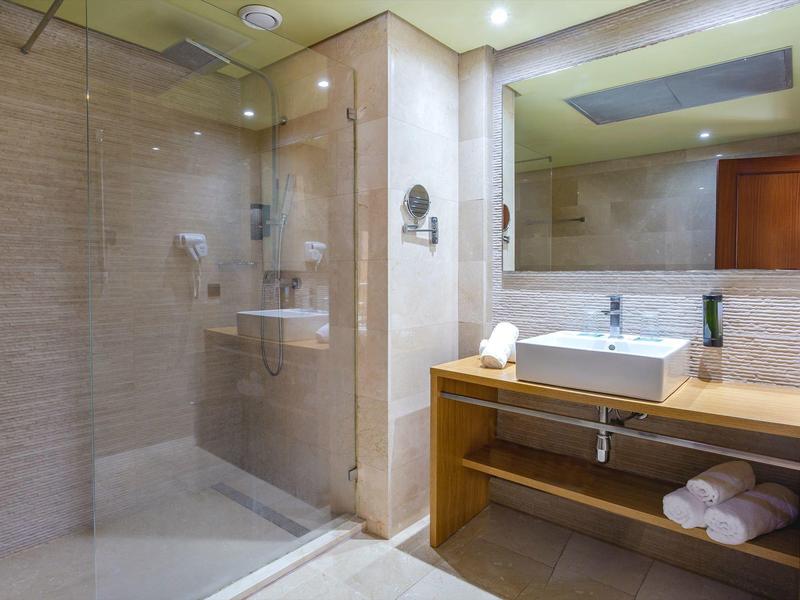 Nowoczesna łazienka z szklaną kabiną prysznicową, białą umywalką na drewnianej szafce i ręcznikami na półce.