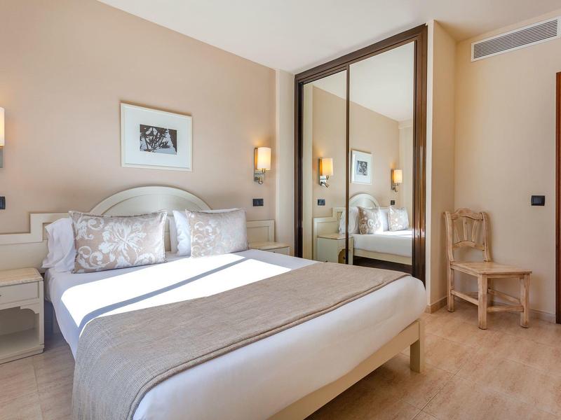 Jasny pokój hotelowy z dużym łóżkiem, stolikami nocnymi, lampami ściennymi, szafą z lustrem i drewnianym krzesłem.