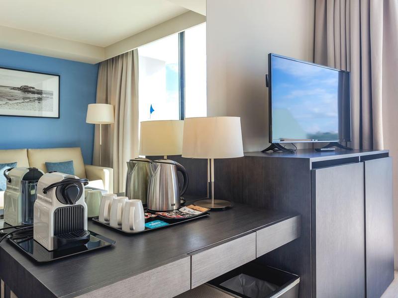 Narożnik pokoju hotelowego z ekspresem do kawy, lampą, telewizorem i łóżkiem przy oknie z zasłonami.