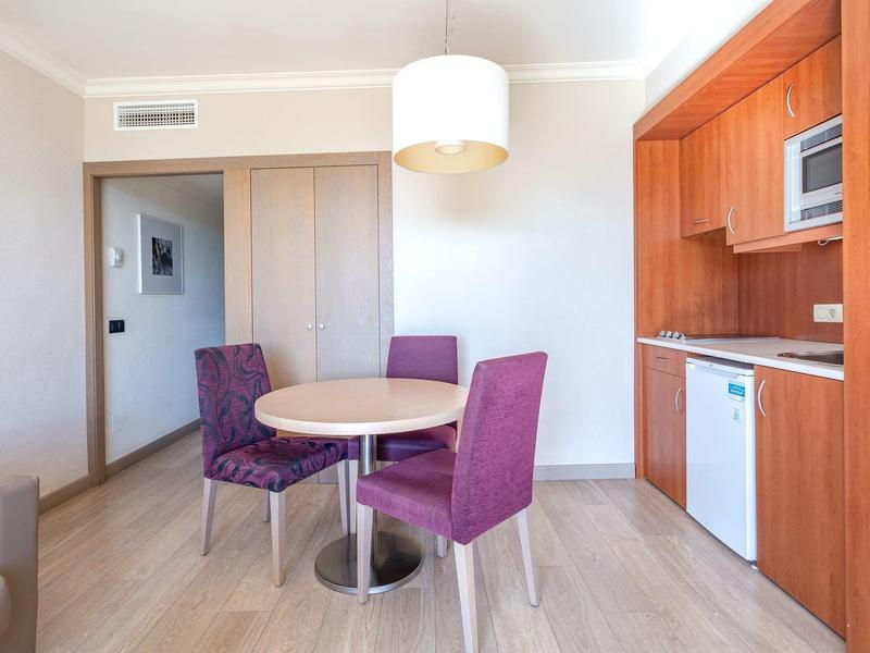 Jasny kąt hotelowy z okrągłym stołem, dwoma fioletowymi krzesłami i drewnianym aneksem kuchennym.