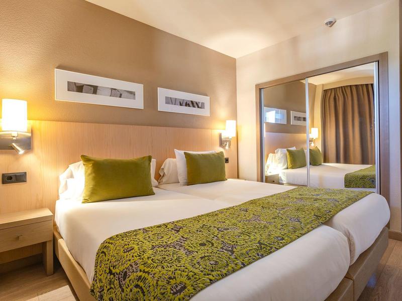 Czysty pokój hotelowy z podwójnym łóżkiem, zielonymi poduszkami, stolikami nocnymi i dużym lustrem.