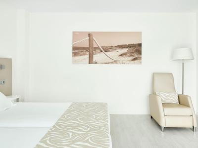 Lichte hotelkamer met twee bedden, een crème ligstoel en minimalistische wanddecoratie.
