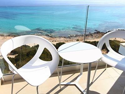 Zwei weiße Stühle und ein kleiner Tisch auf Balkon mit Glasgeländer und Meerblick im Hintergrund.