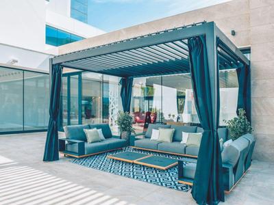 Moderner Pavillon mit blauen Vorhängen, Sofas und Teppich auf heller Terrasse neben Glasfassade.