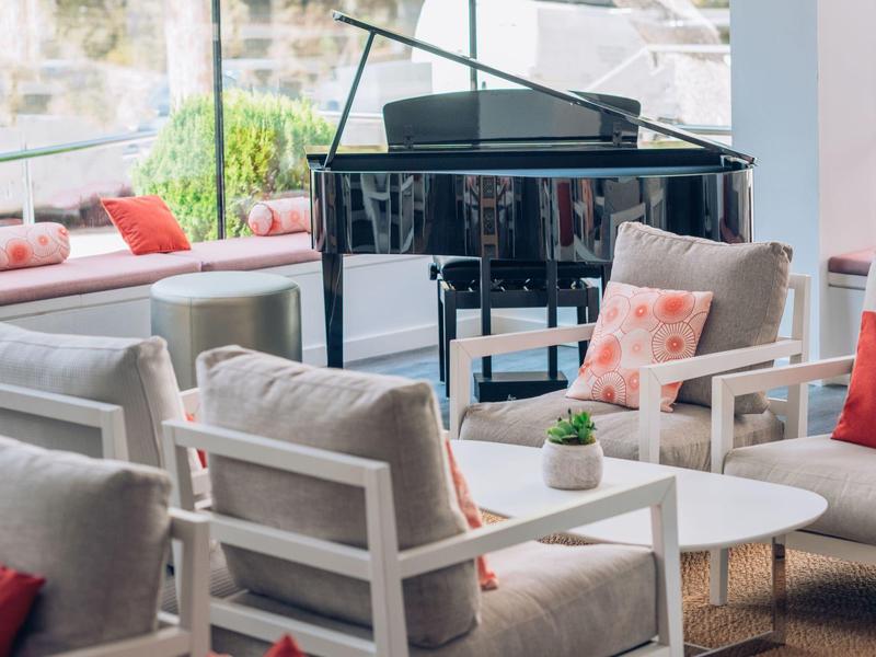 Moderne Lounge mit grauen Sesseln, weißen Tischen, bunten Kissen und schwarzem Flügelklavier.