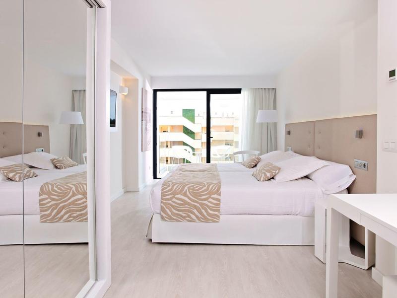 Helles Schlafzimmer mit großem Bett, beiger Bettwäsche, Spiegeln und Balkon mit Glasgeländer.