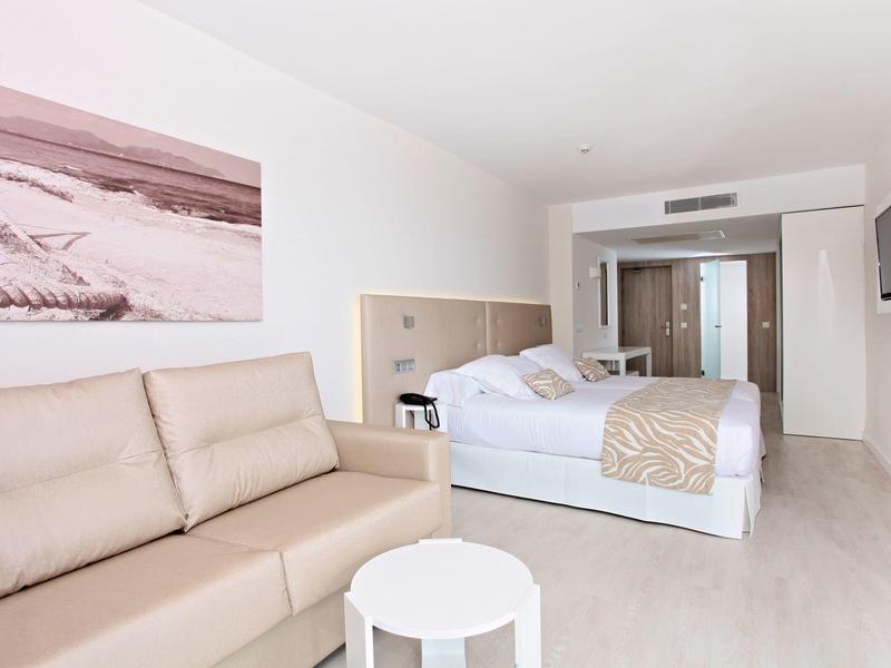 Hell eingerichtetes Hotelzimmer mit großem Bett, Sofa, kleinem runden Tisch und dekorativem Wandbild.