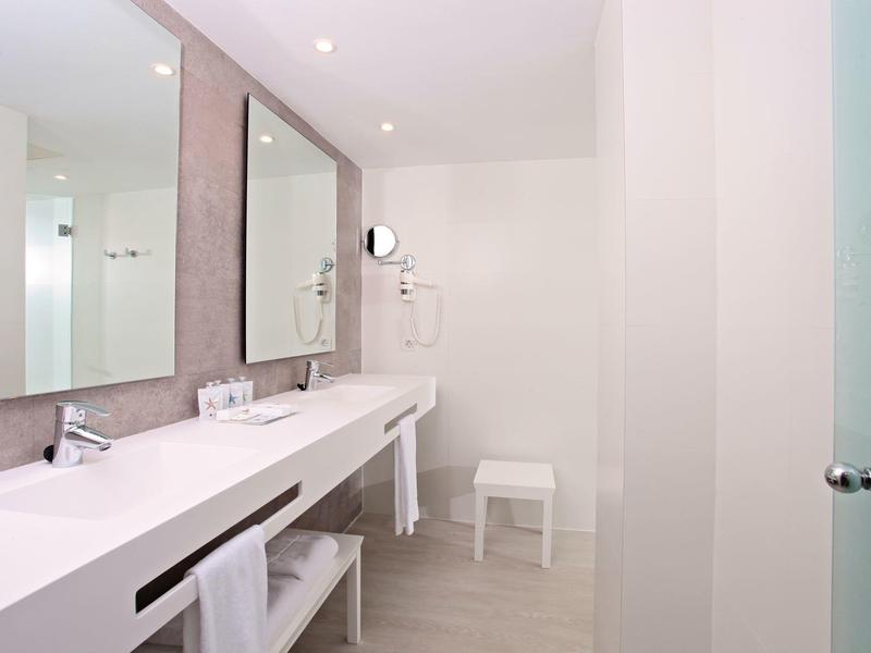 Hell gestaltetes Badezimmer mit weißem Waschbecken, großem Spiegel und Holzhocker unter der Theke.