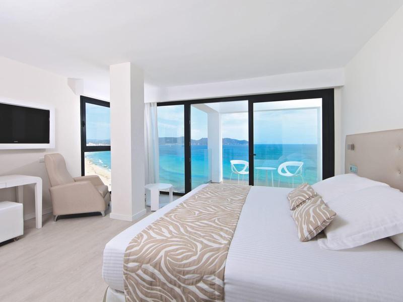 Helles Hotelzimmer mit Doppelbett, Sessel und großer Glasfront mit Meerblick.