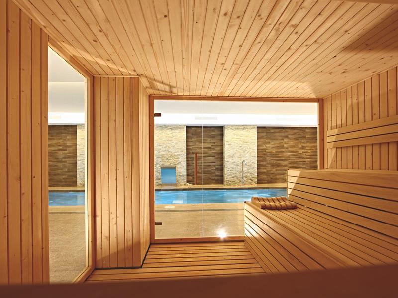 Saunaraum aus hellem Holz mit Glasfenster, Blick auf beheiztes blaues Schwimmbad außen.
