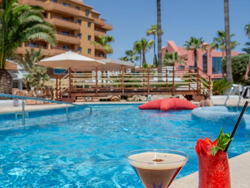 Due cocktail accanto a una piscina di un hotel con palme e cielo azzurro sullo sfondo.
