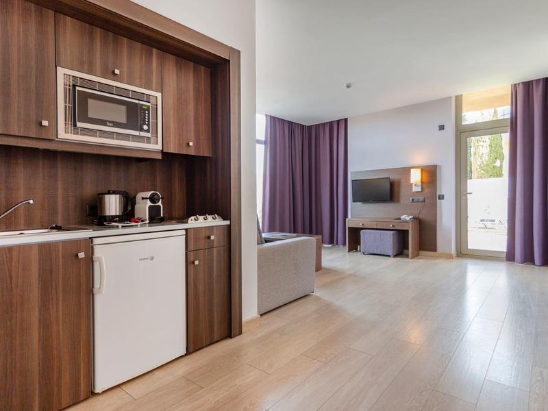 Camera d'hotel luminosa con angolo cottura, TV e grande finestra con tende viola.