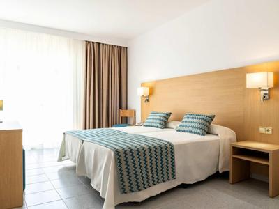 Brightly furnished hotel room with double bed, two nightstands, and large windows with curtains.