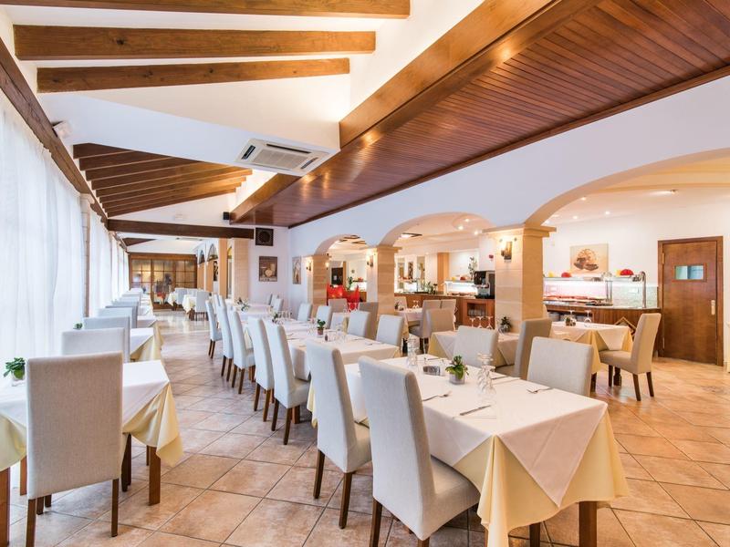 Ampia sala ristorante luminosa con sedie bianche, tavoli apparecchiati e soffitto con travi in legno.