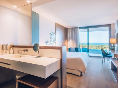 Modern hotel room with a bed, desk, open bathroom, and a sea view through a large window.