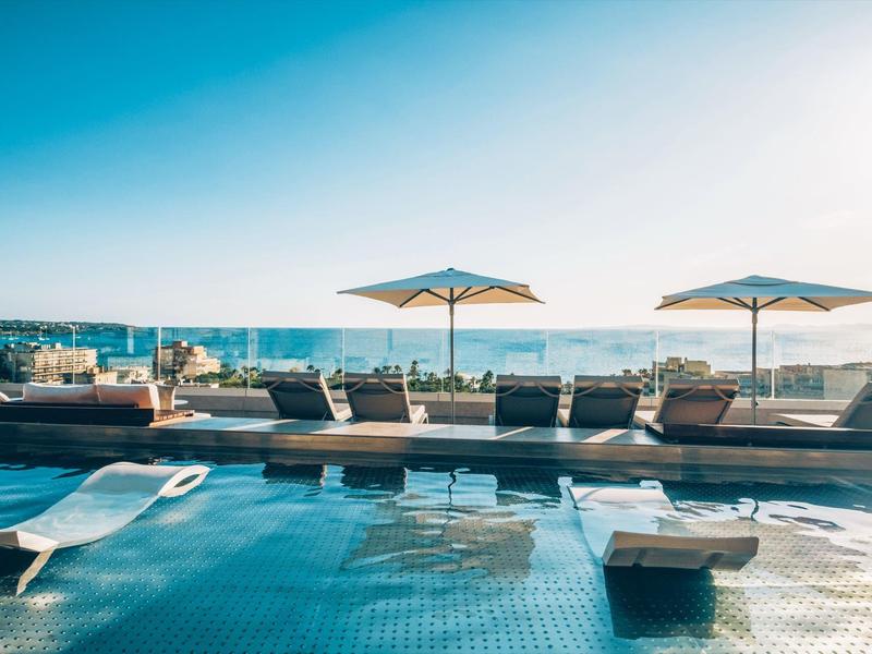 Rooftop pool with lounge chairs and umbrellas overlooking the ocean under a clear sky.