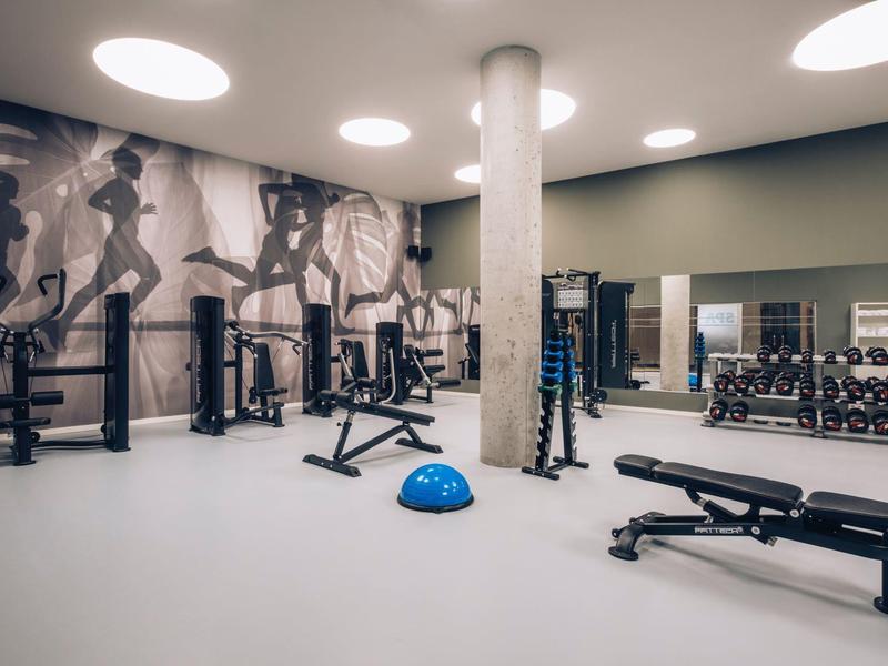 Modern gym with weights, benches, and fitness equipment, featuring large wall mural and round ceiling lights.