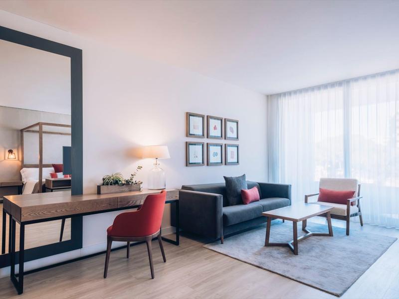 Modern hotel room with sofa, red chairs, large mirror, and natural light through sheer curtains.