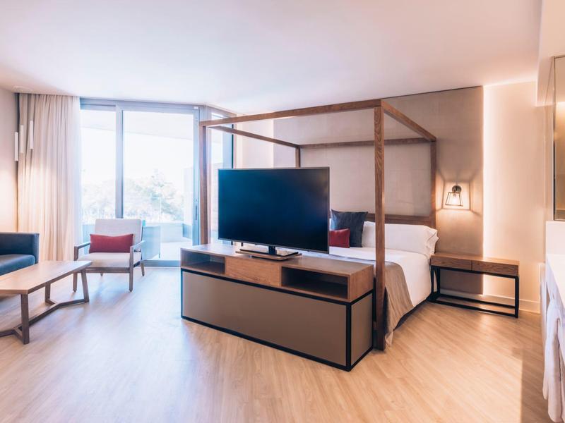 Bright hotel room with large window, double bed, TV, sofas, and wooden floor.