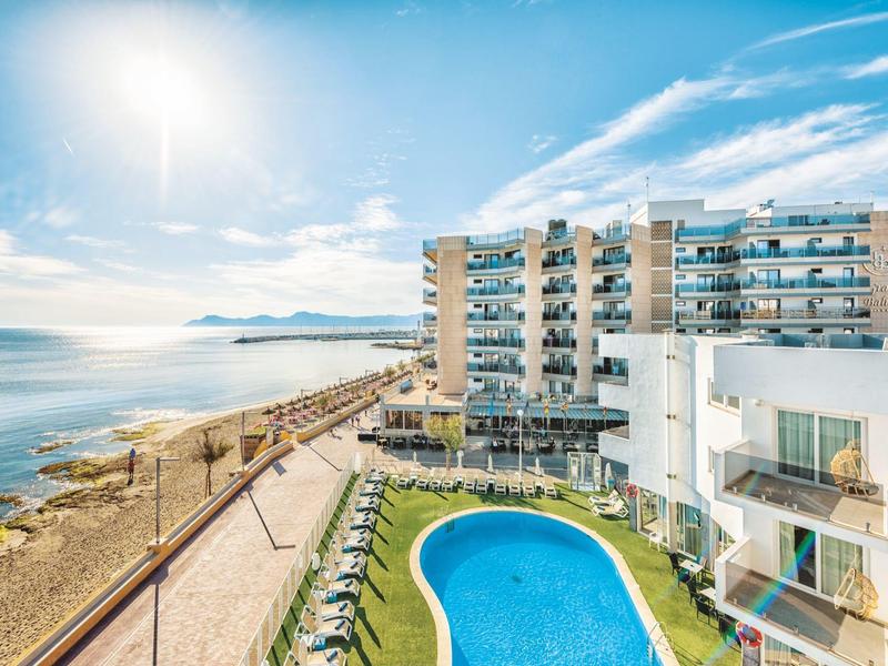 Hotel with outdoor pool next to coastal promenade and sea under blue sky.