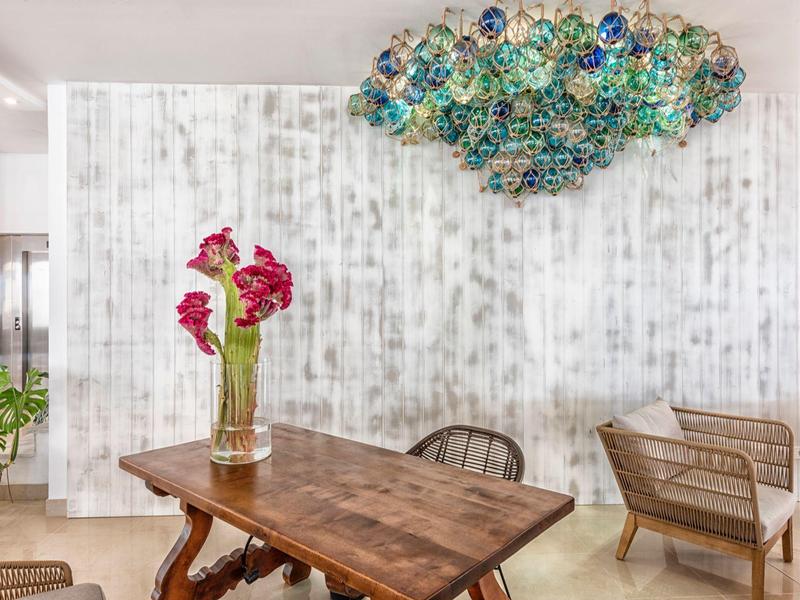 Modern dining area with wooden table, glass vase with red flowers, and colorful ceiling lighting.