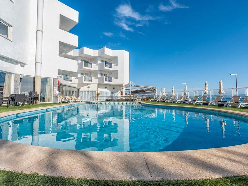 Round pool in front of a modern white hotel with blue sky in the background.