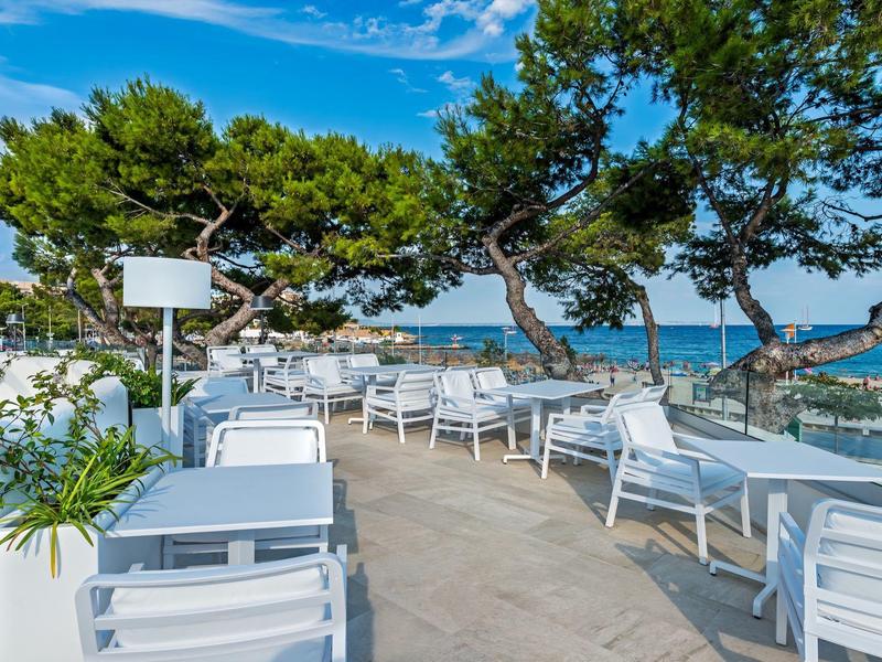 Terrasse extérieure avec tables blanches donnant sur la mer sous un ciel bleu et des arbres verts.