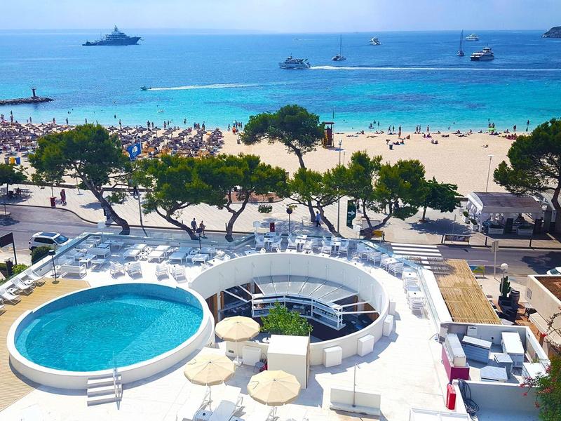 Vue d'une plage animée avec piscine ronde et chaises longues au premier plan.