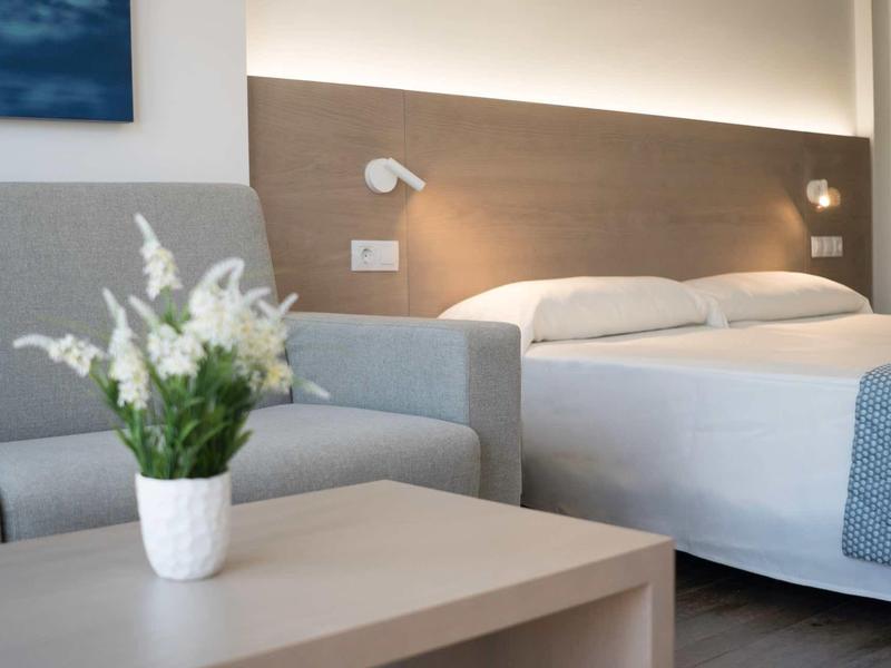 Camera d'hotel moderna con letto matrimoniale, divano grigio e decorazione floreale su un tavolo.