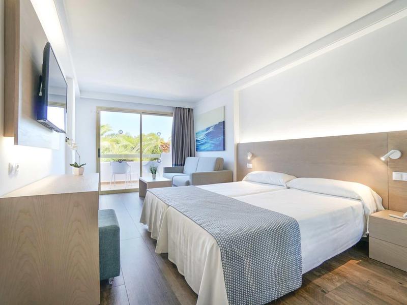 Camera d'hotel moderna con letto matrimoniale, zona salotto e balcone con vista su alberi verdi.