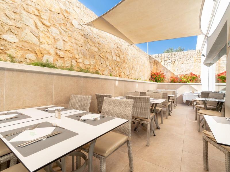 Area esterna moderna di un ristorante con tavoli e sedie tra muri in pietra e vele da sole.