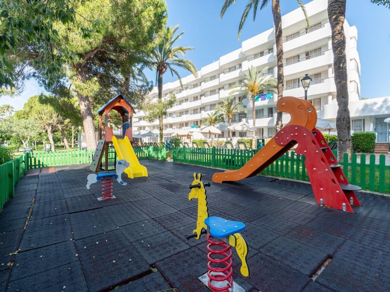 Aree giochi per bambini con altalene e scivoli colorati davanti a un edificio di hotel sotto un cielo blu.