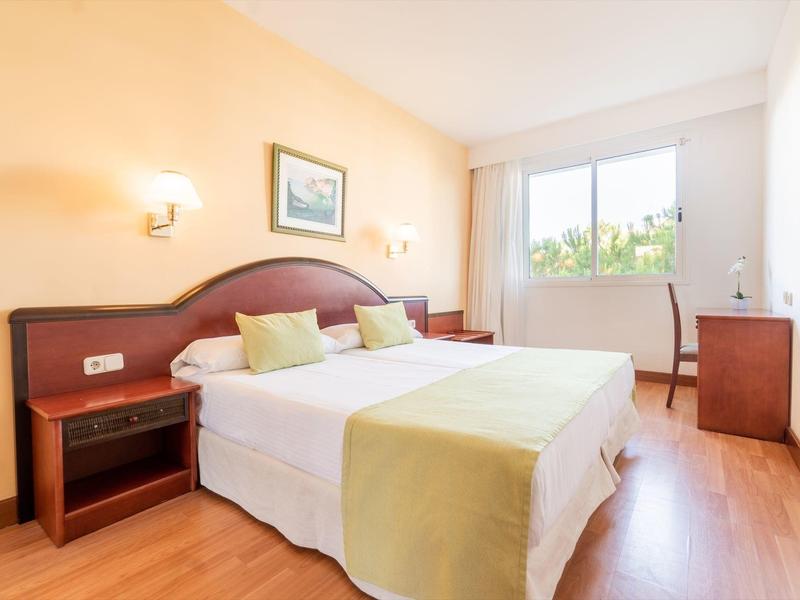 Camera d'hotel luminosa con due letti, pavimento in legno, scrivania e grande finestra con vista esterna.