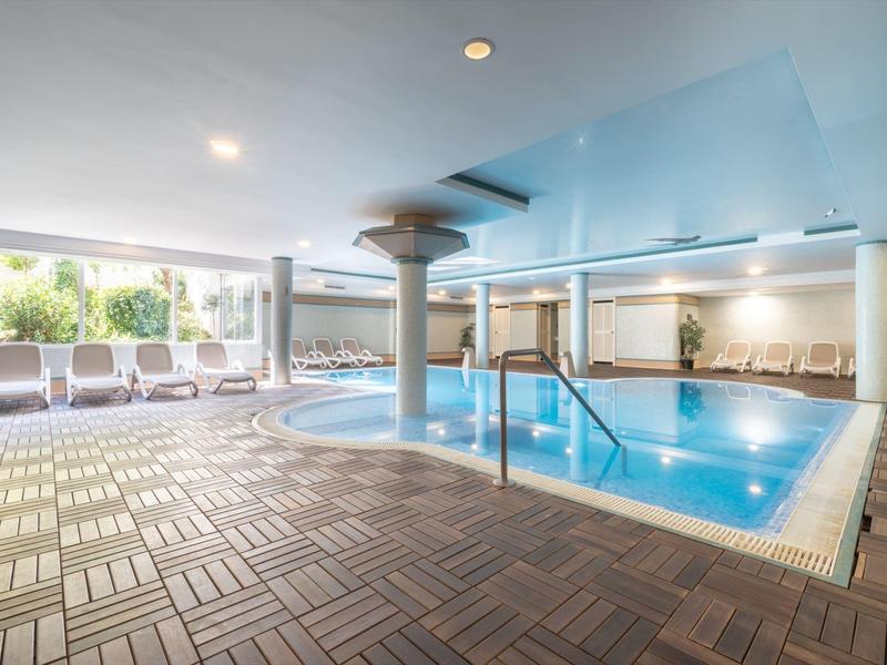 Area piscina coperta con acqua blu, lettini e grandi finestre che lasciano entrare la luce del giorno.