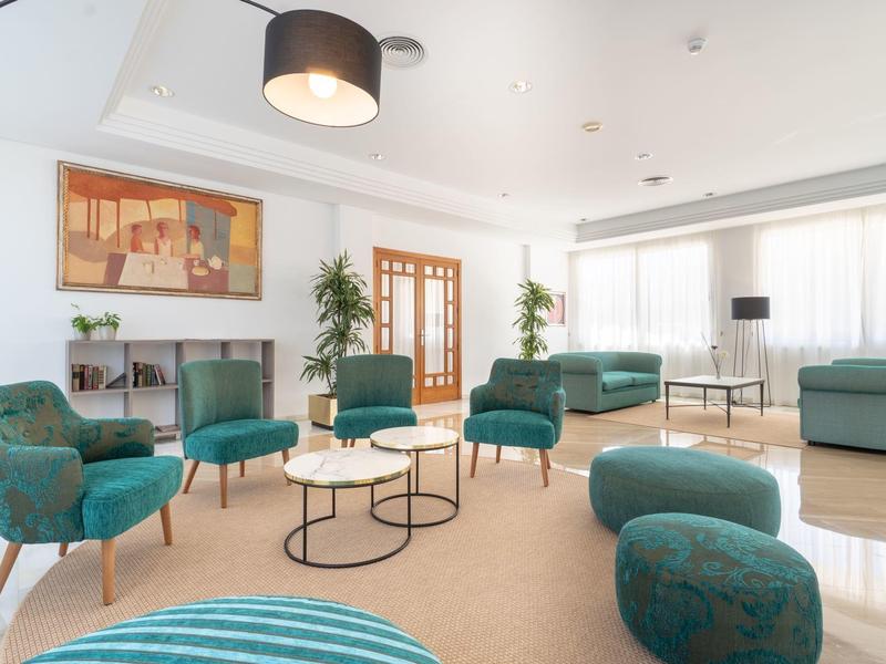 Hall d'hotel luminosa con poltrone turchesi, divani, tavolini da caffè e grande finestra.