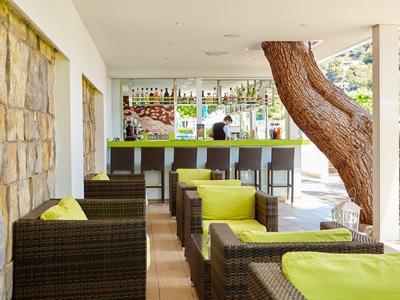 Moderner Außenbereich mit Rattanmöbeln, grünen Sitzkissen und Bar im Hintergrund unter Dach.