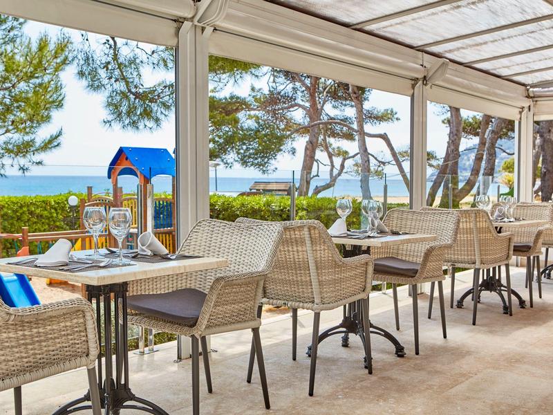 Terrassenrestaurant mit geflochtenen Stühlen, Holztischen und Blick auf Meer und Bäume.
